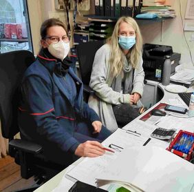 Zwei Frauen sitzen in einem Büro nebeneinander. Links sitz Elena Lützeler von den Maltesern und rechts Gina Raths von der Caritas. Beide schauen in die Kamera und tragen Masken. 